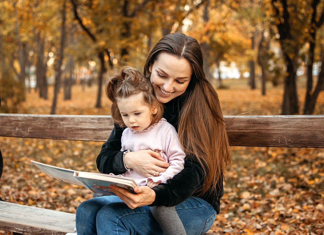 Read Our Reviews - Cheerful Mother Sitting on a Park Bench During the Fall Season with her Daughter on her Lap While Reading a Book